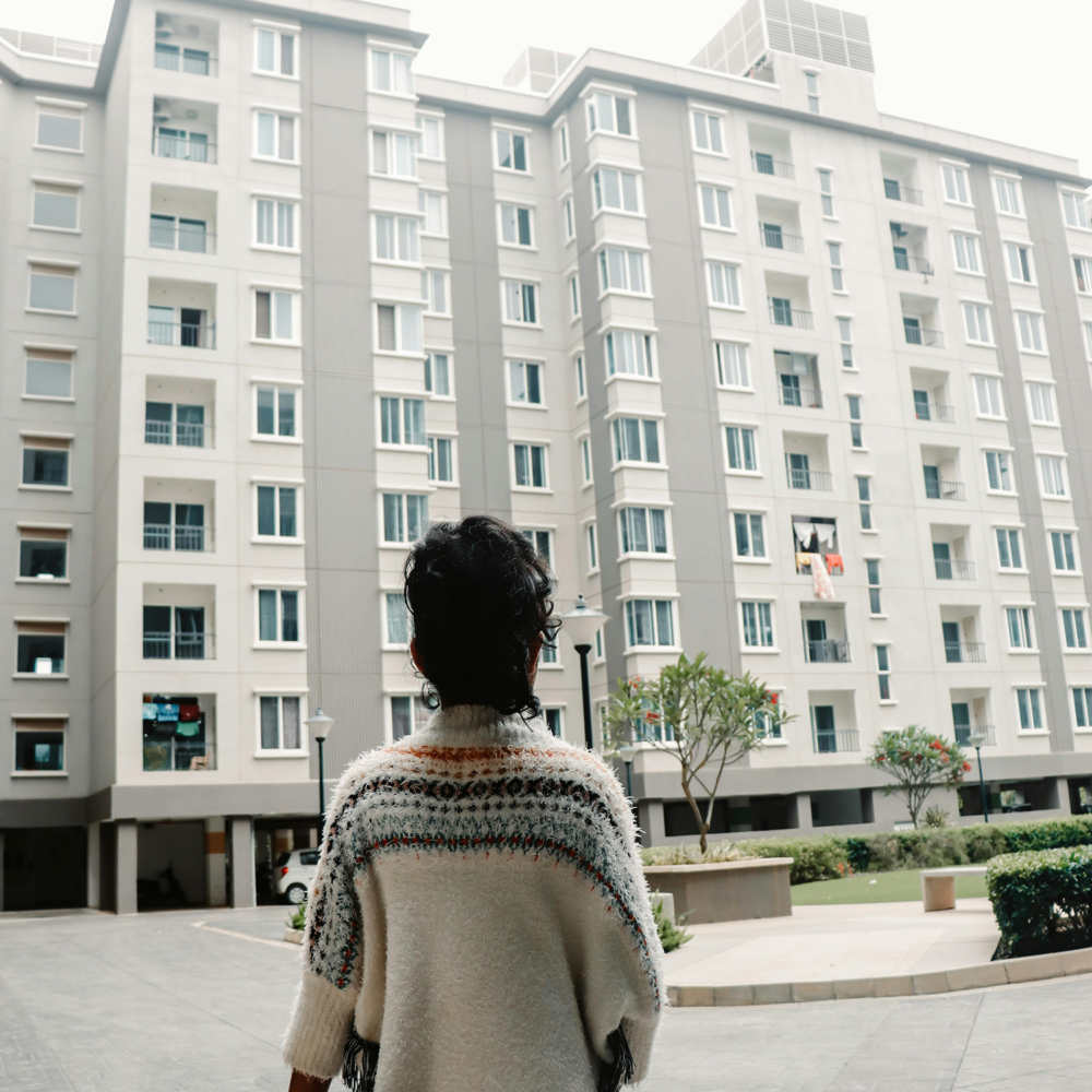 A person with curly hair stands wearing a knitted sweater while looking at a modern multi-story apartment building.